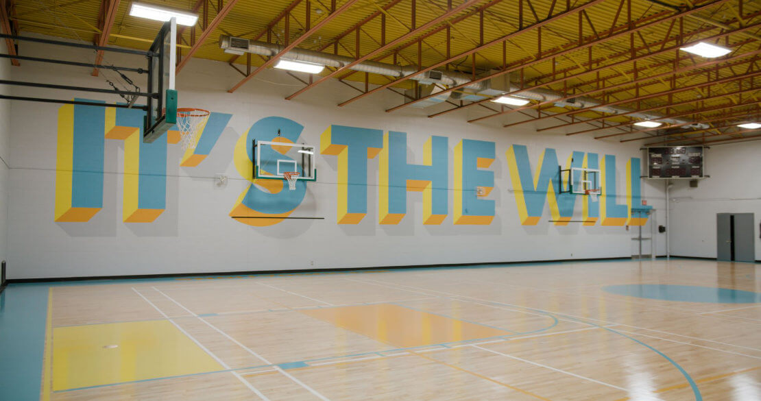 Eine leere Basketballhalle, auf der Wand steht "it's the will"