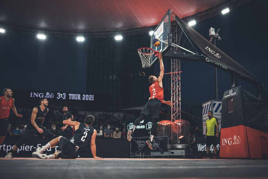 Beim Streetball-Spiel macht ein Spieler einen Korbleger, ein Gegenspieler sitzt auf dem Boden