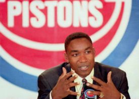 Isiah Thomas im Anzug bei einer Pressekonferenz, dahinter das Logo der Detroit Pistons