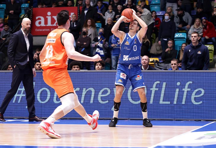 Zwei Basketballspieler und ein Trainer sind auf dem Spiel Feld und im Hintergrund schauen Fans zu