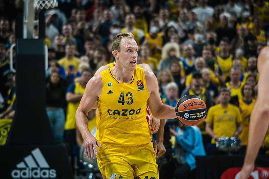 Luke Sikma hält ein Basketball in der linken Hand und im Hintergrund sieht man eine Zuschauermenge sitzen.