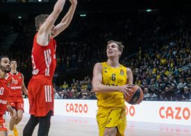 Auf der linken Seite springt ein Basketballspieler im roten Trikot hoch und auf der rechten seite ist ein Basketballspieler im gelben Trikot der den Basketball mit beiden Händen festhält