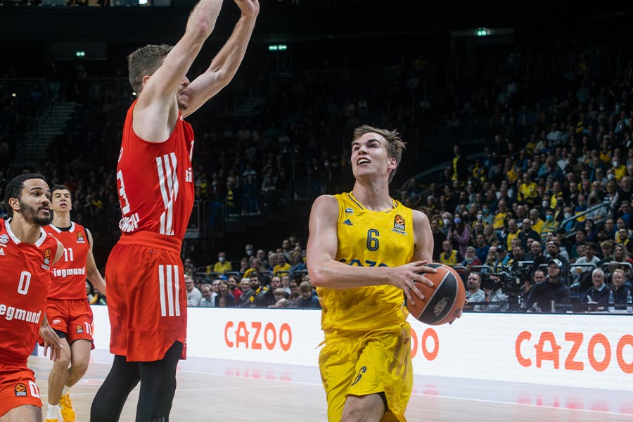 Auf der linken Seite springt ein Basketballspieler im roten Trikot hoch und auf der rechten seite ist ein Basketballspieler im gelben Trikot der den Basketball mit beiden Händen festhält
