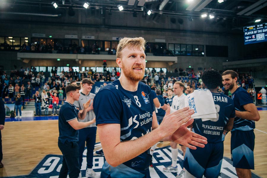 Der Basketballspieler Max Ugrai von der Mannschaft MPL Academics Heidelberg klatscht in seine Hände und im Hintergrund sieht man seine Mannschaft