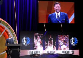 Dirk Nowitzki an einem Redepult bei seiner Aufnahme in die Hall of Fame, rechts neben ihm Bilder aus seiner Karriere