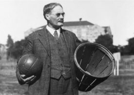 Dr. James Naismith mit einem Ball in der rechten und einem Korb in der linken Hand