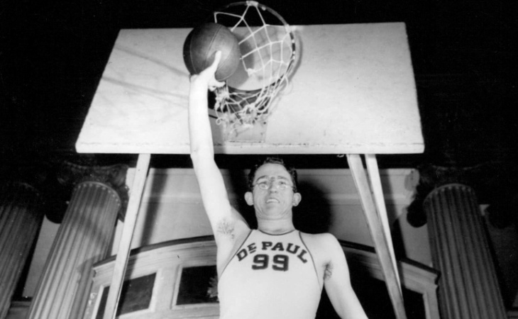 George Mikan hält einen Ball mit seinem ausgestreckten rechten Arm knapp unter einen Korb